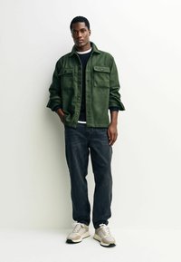 Grüne Wollmischjacke mit zwei Brusttaschen, navyblauer strukturierter Pullover, dunkle Jeans und beige Sneakers mit weißen Schnürsenkeln.