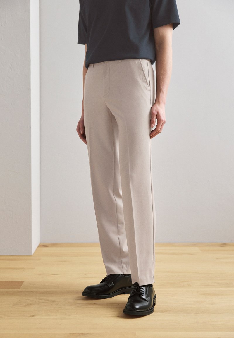 Pantaloni beige su misura con design a gamba dritta, tessuto leggero e tasche laterali, abbinati a scarpe eleganti nere e una maglietta blu navy.