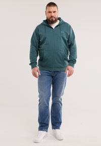 Szakállas férfi, aki tengerzöld cipzáras kapucnis pulóvert, kék farmert és fehér tornacipőt visel, és egy egyszínű világos háttér előtt áll.