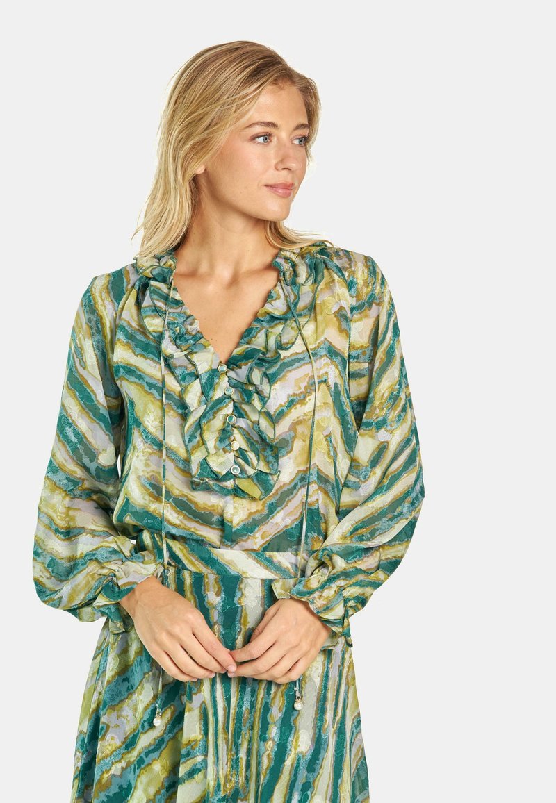 Blusa verde e gialla con fantasia, colletto arricciato e bottoni. Maniche lunghe con polsini arricciati, realizzata in tessuto leggero.