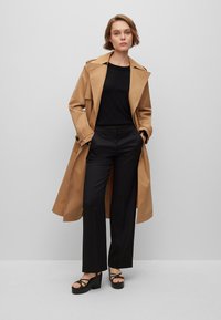 Beige trenchcoat over en svart topp, kombinert med svarte vide bukser og chunky svarte platåsandaler. Glatt stoff, klassisk design.
