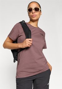 Bordeaux katoenen t-shirt met een ronde halslijn en een zwart logo. Gecombineerd met zwarte broeken en een zwarte rugzak, geaccentueerd met lichte zonnebril.