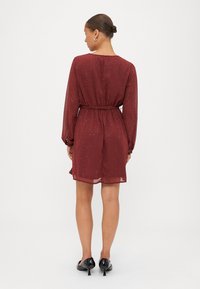 Maroon chiffon haljina s dugim rukavima, pojasom u struku i raspršenim sjajnim detaljima. Duljine do sredine butina s glatkom, lepršavom teksturom. Crne cipele.