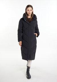 Langer schwarzer Puffer-Mantel mit Kapuze, ausgestattet mit einem Chevron-gequilteten Muster, zwei seitlichen Taschen und einer glatten Oberfläche. Das Model trägt dunkle Stiefel und helle Hosen.