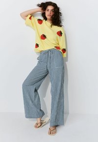 Geel trui met rode aardbeienpatronen, gecombineerd met blauw-witte geruite wijde broek, gedragen met beige slippers.
