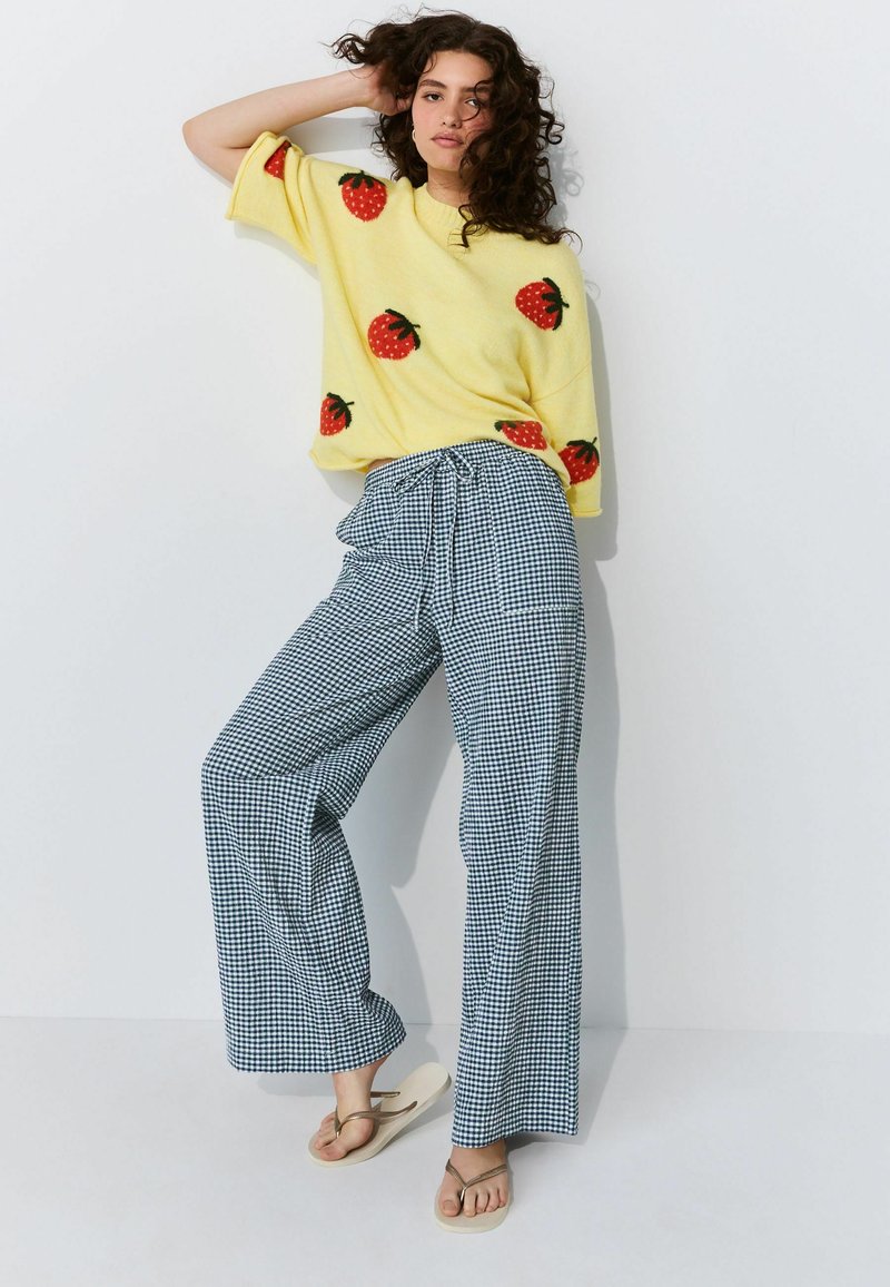 Geel trui met rode aardbeienpatronen, gecombineerd met blauw-witte geruite wijde broek, gedragen met beige slippers.