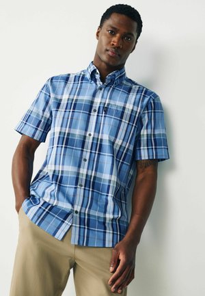 Junger Mann trägt ein blaues, kariertes Hemd mit kurzen Ärmeln und beige Hosen, steht mit einer Hand in der Tasche vor einer weißen Wand.