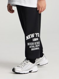 Czarne spodnie dresowe z białym napisem "NEW YORK 1984 HIGH SCHOOL ATHL.DEPT ORIGINAL" przy kostce, zestawione ze srebrnymi butami sportowymi.