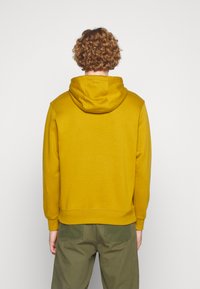 Sweatshirt mostarda com capuz ajustável, punhos e barra canelados. O tecido parece liso, combinado com calças verde-oliva.