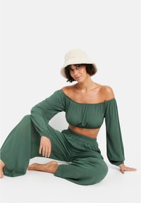 Zeleno dvojnoslojno oblačilo, ki vključuje crop top brez naramnic z dolgimi rokavi in ohlapne hlače z visokim pasom. Teksturo tkanine z gladko površino.