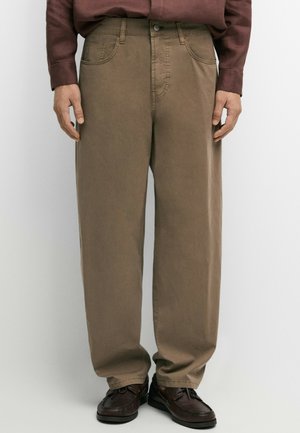 Persona che indossa pantaloni marroni a taglio largo e scarpe marrone scuro, in piedi davanti a uno sfondo chiaro e uniforme con le mani rilassate ai lati.