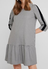 Černobílé houndstooth šaty s tříčtvrtečními rukávy, nařaseným lemem a kontrastními černými bočními panely. Měkká textilie.