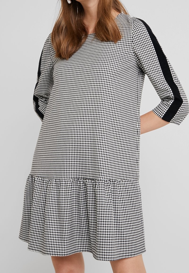 Černobílé houndstooth šaty s tříčtvrtečními rukávy, nařaseným lemem a kontrastními černými bočními panely. Měkká textilie.