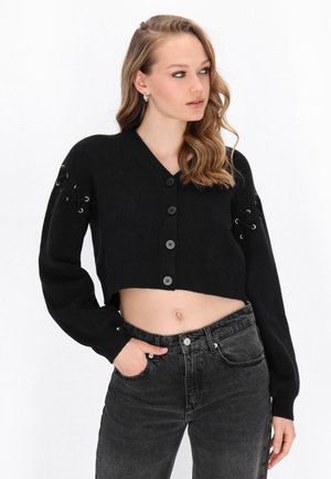 Kvinde iført en sort kort cardigan med knapper og snøredetaljer på ærmerne, kombineret med højtaljede mørkegrå jeans.