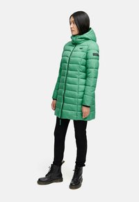 Groene gewatteerde jas met een capuchon, voorzien van een rits aan de voorkant, een gequilt ontwerp en zwarte accenten. Draagt met zwarte broeken en veterlaarzen.