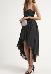 Femme portant une robe noire sans bretelles à ourlet asymétrique, des talons noirs à bride de cheville, tenant une pochette argentée sur fond clair.