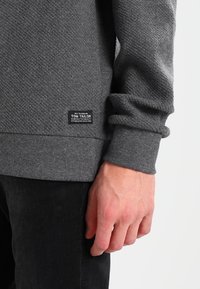 Sudadera gris texturizada con puños y dobladillo acanalados. Presenta una etiqueta negra con "TOM TAILOR" en la parte inferior izquierda. Pantalones negros son parcialmente visibles.