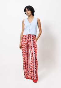 Helles, hellblaues, ärmelloses Rippoberteil mit Knöpfen vorne, kombiniert mit hochsitztenden, bunten, gemusterten weiten Hosen. Rote Schuhe runden das Outfit ab.