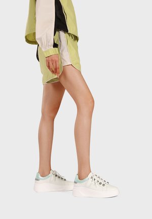 Persona con indosso pantaloncini sportivi verde chiaro e beige e giacca, sneakers con zeppa bianca con dettagli azzurri, in piedi davanti a uno sfondo neutro.