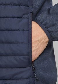 Navy puffer jacket detail showing a textured zip pocket and cuff. Material is quilted with a smooth finish; zipper has a black pull tab.