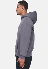 Grå hætte sweatshirt med en frontlomme, løs pasform og ribbede manchetter. Har et logo på venstre ærme. Glat stoftekstur.