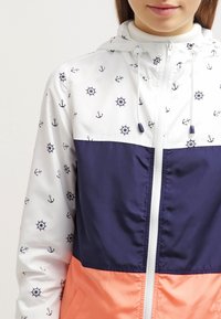 Farverig vindjakke med en hvid overdel med navy anker- og hjulmønstre, navy midtersektion og koralfarvet nederste sektion. Med hætte og lynlås.