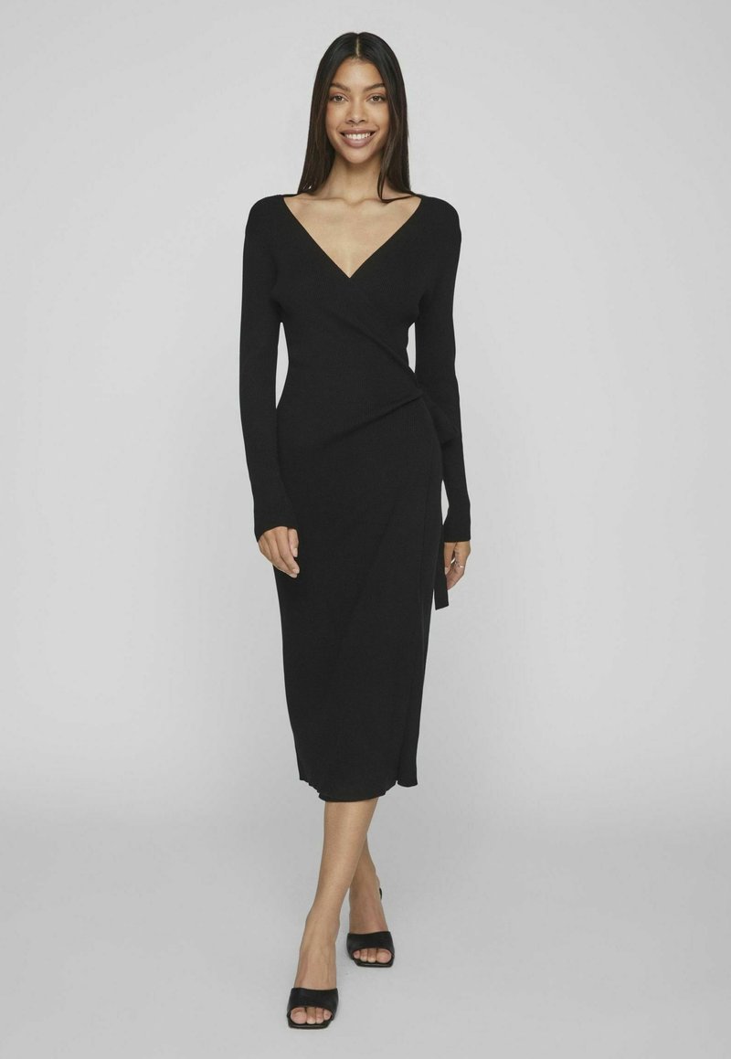 Vestido negro de manga larga con cinturilla en material de canalé, con un escote profundo en V y longitud hasta los tobillos. Combinado con tacones destalonados negros.