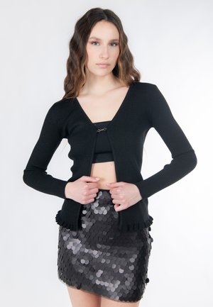 Schwarzer, gerippter Cardigan mit langen Ärmeln und V-Ausschnitt, vorne mit einem Verschluss. Kombiniert mit einem glänzenden schwarzen Paillettenrock.
