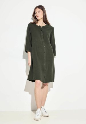 Robe verte olive à boutons en tissu texturé avec un col rond et des manches trois-quarts, associée à des baskets blanches.