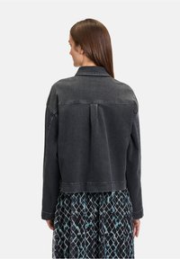 Czarna dżinsowa kurtka o luźnym kroju, z plisą z tyłu, spiczastym kołnierzykiem i kontrastowym przeszyciem. Noszona na drukowanej sukience.