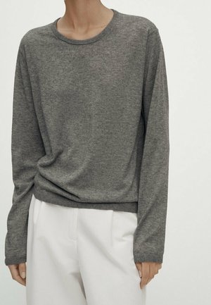 Personne portant un pull gris uni à manches longues et un pantalon blanc, debout sur un fond clair uni.
