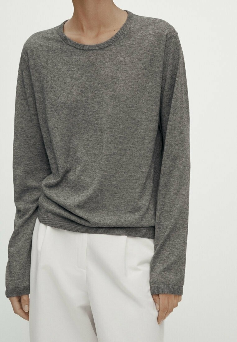 Personne portant un pull gris uni à manches longues et un pantalon blanc, debout sur un fond clair uni.