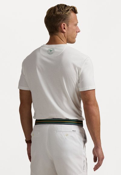 Weißes T-Shirt mit kurzen Ärmeln und einem runden Logo-Detail auf der Rückseite. Getragen mit hellen Hosen, die einen mehrfarbigen Gürtel haben.