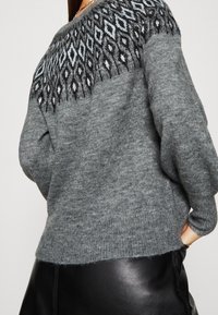 Femme portant un pull gris avec un motif en losanges noir et blanc sur l'empiècement et une jupe en cuir noir, la main dans la poche.