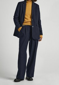 Person wearing navy pinstripe suit with wide-leg trousers, mustard knit sweater, and black chunky shoes, standing against a plain background.