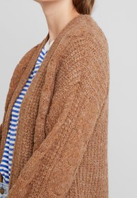Brown knitted cardigan with a textured pattern, open front, and long sleeves. Worn over a blue and white striped shirt.
