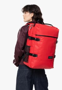 Zaino rosso realizzato in tessuto resistente, dotato di cinghie laterali regolabili, maniglia superiore e tasche laterali in rete. Design rettangolare con dettagli neri.