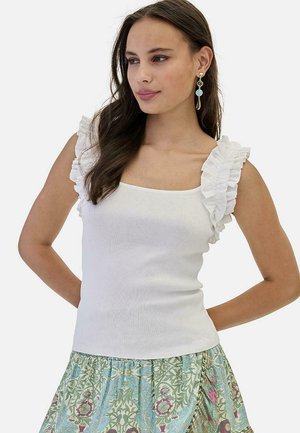 Mujer con cabello largo y oscuro, vistiendo una blusa sin mangas blanca con volantes y una falda floral azul verdosa, complementada con pendientes colgantes.