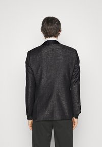 Černý blazer s texturovaným povrchem, hedvábnými klopami a dvěma knoflíky, s dokonale padnoucím střihem a hladkým podšíváním, nošený přes tmavé kalhoty.