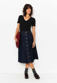 Jupe en denim en forme de A avec des boutons sur le devant et une taille haute, associée à un haut noir à col en V. Les accessoires comprennent un sac rouge à franges et des chaussettes grises.