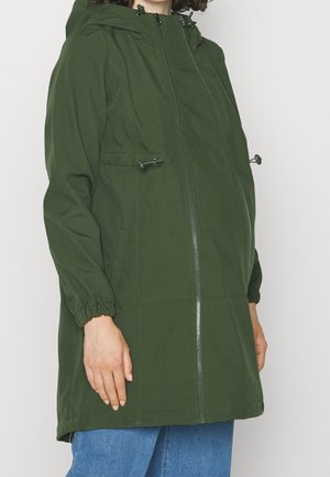 Persoon draagt een lange groene jas met capuchon, ritssluiting aan de voorkant en verstelbare trekkoorden, gecombineerd met blauwe spijkerbroek, staand tegen een witte achtergrond.