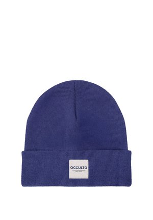 Junger Mann mit blondem Haar, der eine blaue Beanie und ein rotes kariertes Hemd trägt, schaut mit neutralem Gesichtsausdruck auf weißem Hintergrund geradeaus.