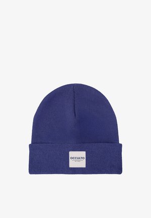 Junger Mann mit blondem Haar, der eine blaue Beanie und ein rotes kariertes Hemd trägt, schaut mit neutralem Gesichtsausdruck auf weißem Hintergrund geradeaus.