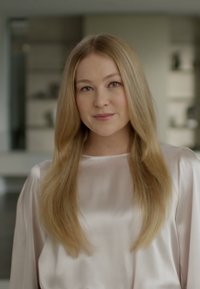 Jasnoróżowa satynowa bluzka z długimi rękawami, noszona przez osobę z długimi, prostymi blond włosami. Gładka faktura, minimalistyczny design, delikatny wygląd.