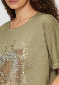 Olivgrünes T-Shirt mit weitem Ausschnitt, das ein metallisches Blumenmuster in Bronze- und Silbertönen zeigt, mit strukturiertem Stoff und kurzen Ärmeln.
