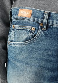Blauwe denim jeans met een vervaagd uiterlijk, voorzien van een leren label met de tekst "REPLAY", koperen knopen en een klassieke vijf-pocket ontwerp.