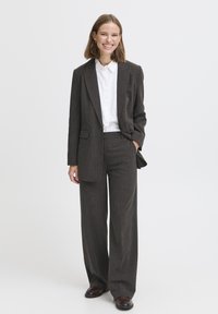 Glimlachende vrouw met schouderlang haar, gekleed in een donkergrijs pak, witte overhemden en bruine schoenen, staand met één hand in de zak.