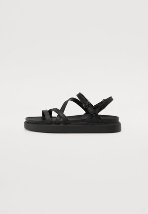 Sort flad sandal med tre tynde remme over foden og en ankelrem med spænde på en hvid baggrund.