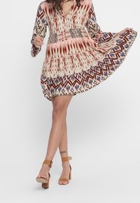 Vestido de manga larga con escote en V en color crema, decorado con patrones tribales multicolores. Presenta una falda acampanada y tres volantes en capas, complementados con sandalias de tacón.
