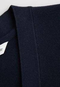 Szczegół granatowego swetra dzianinowego, pokazujący grubą ramiączko i teksturowany materiał. Widoczna metka wskazująca markę. Materiał wydaje się miękki i rozciągliwy.